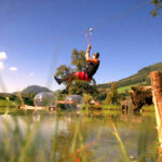 Eine Person in einem Gurtzeug fährt mit einer Seilrutsche über einen Teich, während eine andere Person auf einer Holzplattform steht. Im Hintergrund sind grüne Hügel und Bäume zu sehen.