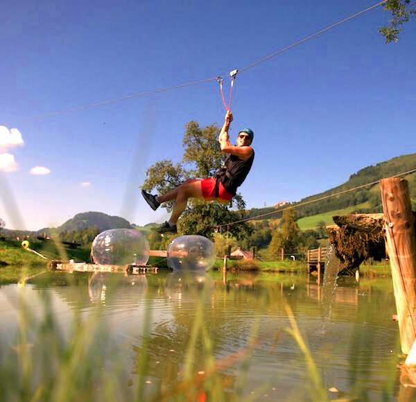 Eine Person in einem Gurtzeug fährt mit einer Seilrutsche über einen Teich, während eine andere Person auf einer Holzplattform steht. Im Hintergrund sind grüne Hügel und Bäume zu sehen.