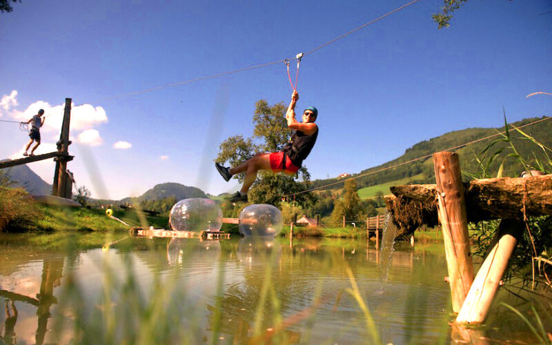 Eine Person in einem Gurtzeug fährt mit einer Seilrutsche über einen Teich, während eine andere Person auf einer Holzplattform steht. Im Hintergrund sind grüne Hügel und Bäume zu sehen.