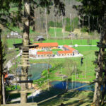 Ein Hochseilgarten in den Baumkronen mit Holzplattformen und Seilen bietet Aussicht auf eine ländliche Landschaft mit Gebäuden, Feldern und einem kleinen Teich darunter.