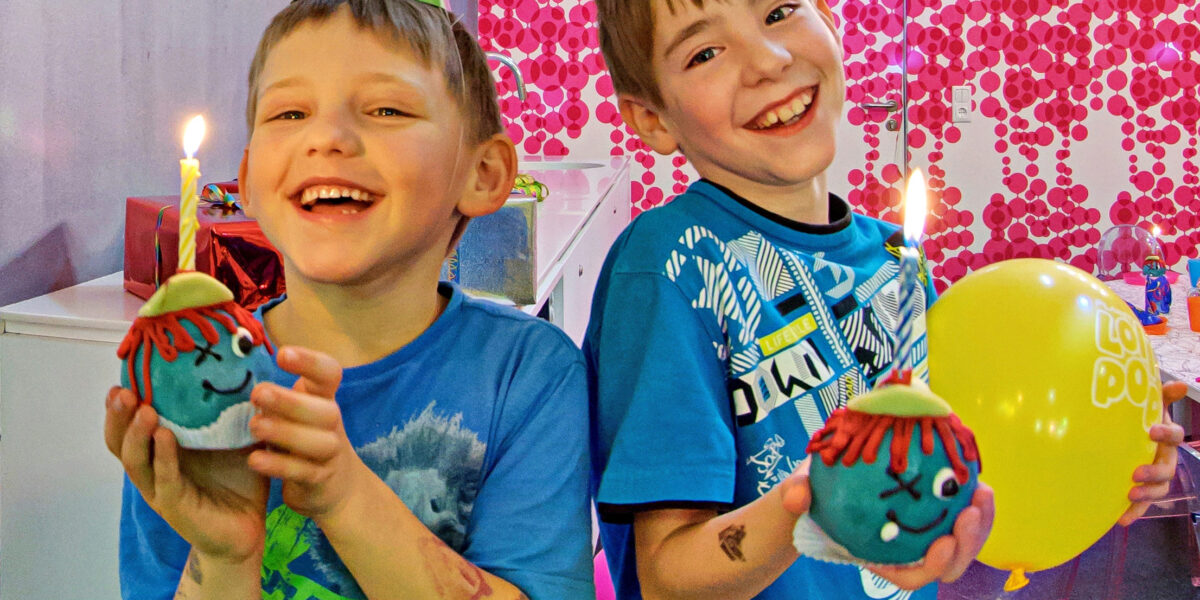 Zwei lächelnde Jungen mit Partyhüten halten auf einer Geburtstagsparty dekorierte Cupcakes mit brennenden Kerzen und einem gelben Luftballon.
