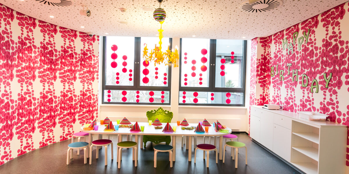 Ein bunt geschmückter Raum mit einem langen, für eine Party gedeckten Tisch, bunten Wandmustern, einem gelben Kronleuchter und einem "Happy Birthday"-Banner am Fenster.