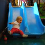 Ein kleines Kind in roter Hose und weißem Hemd rutscht eine blaue Doppelrutsche in einem Indoor-Spielplatz hinunter, während in der Nähe ein paar Plastikbälle auf dem Boden liegen.