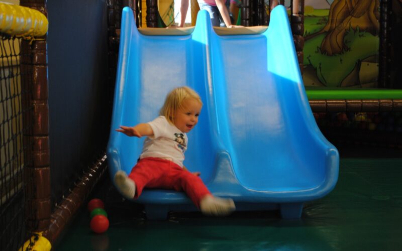Ein kleines Kind in roter Hose und weißem Hemd rutscht eine blaue Doppelrutsche in einem Indoor-Spielplatz hinunter, während in der Nähe ein paar Plastikbälle auf dem Boden liegen.