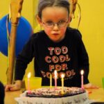 Junges Mädchen mit Brille bläst Kerzen auf einer Geburtstagstorte aus, sie trägt ein "Too Cool for School"-Shirt, im Hintergrund sind Luftballons und Partydekorationen zu sehen.