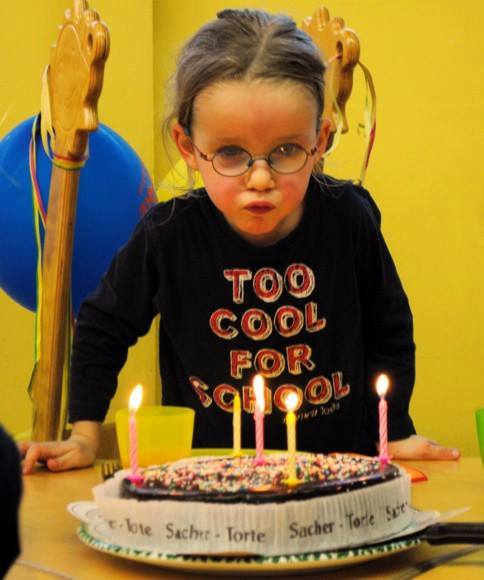 Junges Mädchen mit Brille bläst Kerzen auf einer Geburtstagstorte aus, sie trägt ein "Too Cool for School"-Shirt, im Hintergrund sind Luftballons und Partydekorationen zu sehen.