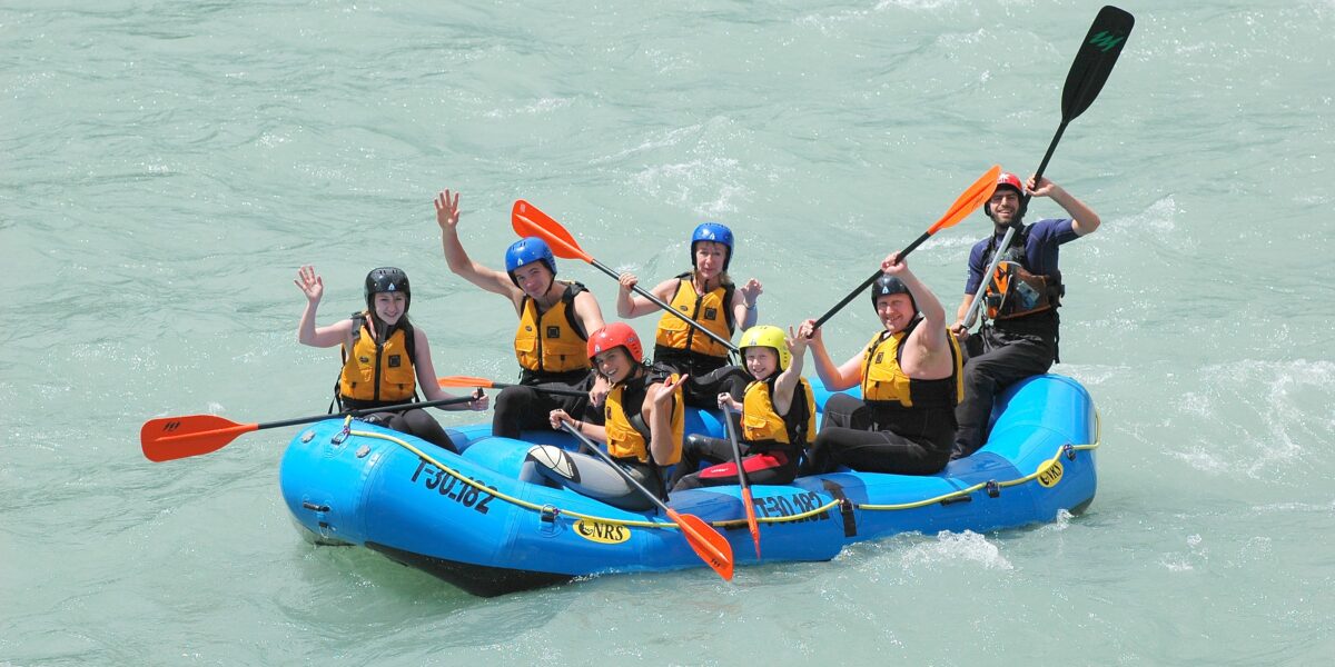 Sechs Personen mit Schwimmwesten und Helmen fahren in einem blauen Schlauchboot auf einem Fluss und winken mit den Paddeln.