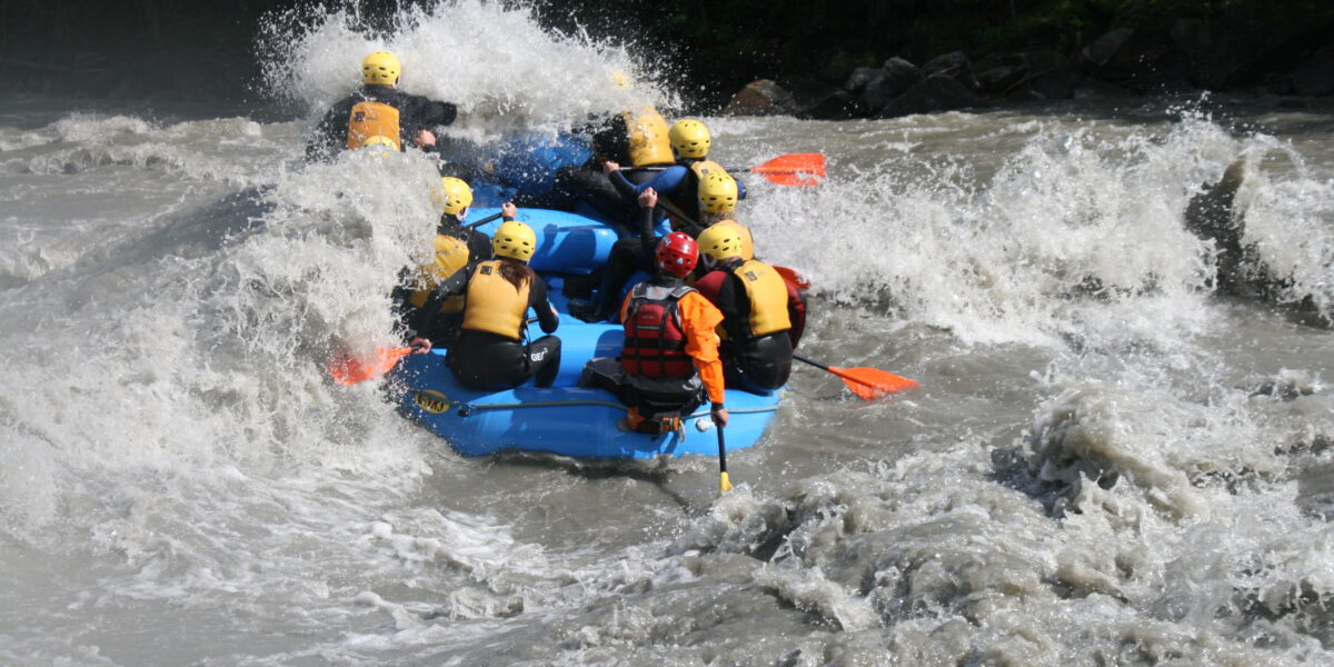 Eine Gruppe von Personen mit Helmen und Schwimmwesten paddelt auf einem blauen Floß durch raue Wildwasser-Stromschnellen.