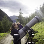 Eine Person steht auf einem unbefestigten Weg in einer bergigen, bewaldeten Gegend und blickt durch ein Fernglas mit einem Spektiv auf einem Stativ in der Nähe.