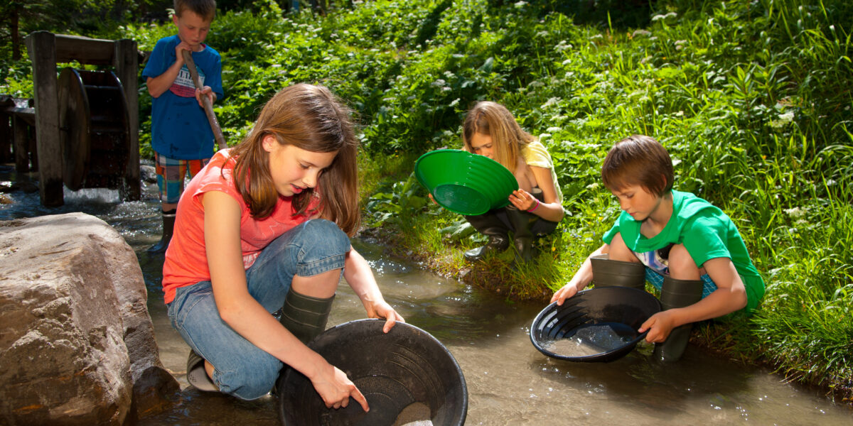 Vier Kinder hocken an einem flachen Bach und suchen an einem sonnigen Tag, umgeben von Grün, mit großen Pfannen nach Gegenständen im Wasser.