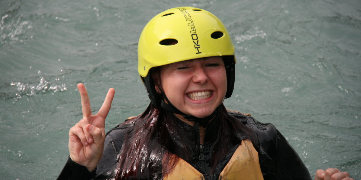 Eine Person mit einem gelben Helm und einer Schwimmweste steht im Wasser, lächelt breit und hält mit einer Hand ein Friedenszeichen hoch.