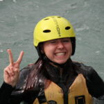 Eine Person mit einem gelben Helm und einer Schwimmweste steht im Wasser, lächelt breit und hält mit einer Hand ein Friedenszeichen hoch.