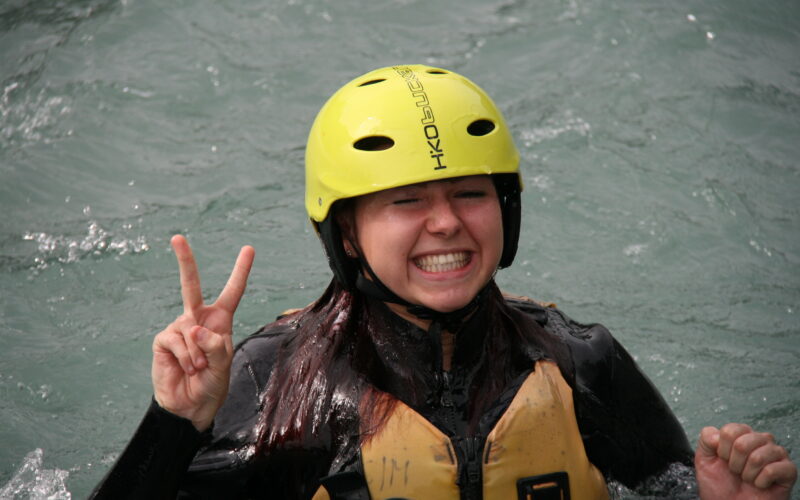 Eine Person mit einem gelben Helm und einer Schwimmweste steht im Wasser, lächelt breit und hält mit einer Hand ein Friedenszeichen hoch.