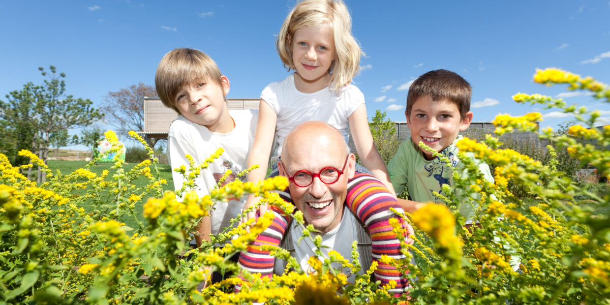 Ein Mann mit roter Brille und drei Kinder lächeln im Freien auf einem Feld mit gelben Blumen unter einem klaren blauen Himmel.