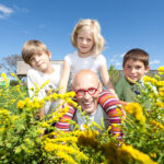 Ein Mann mit roter Brille und drei Kinder lächeln im Freien auf einem Feld mit gelben Blumen unter einem klaren blauen Himmel.