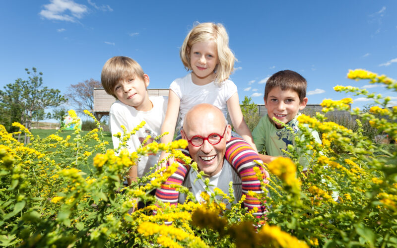 Ein Mann mit roter Brille und drei Kinder lächeln im Freien auf einem Feld mit gelben Blumen unter einem klaren blauen Himmel.
