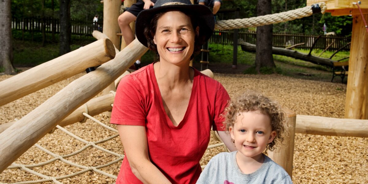 Eine Frau und zwei Kinder sitzen auf einem hölzernen Spielgerät. Im Hintergrund spielen zwei weitere Kinder in einem Baumhaus. Die Szene spielt auf einem bewaldeten Spielplatz im Freien.