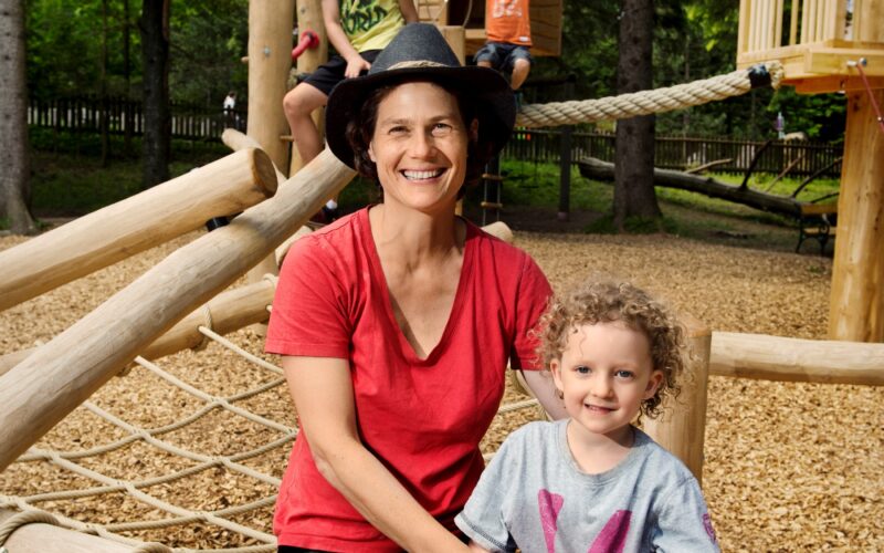 Eine Frau und zwei Kinder sitzen auf einem hölzernen Spielgerät. Im Hintergrund spielen zwei weitere Kinder in einem Baumhaus. Die Szene spielt auf einem bewaldeten Spielplatz im Freien.