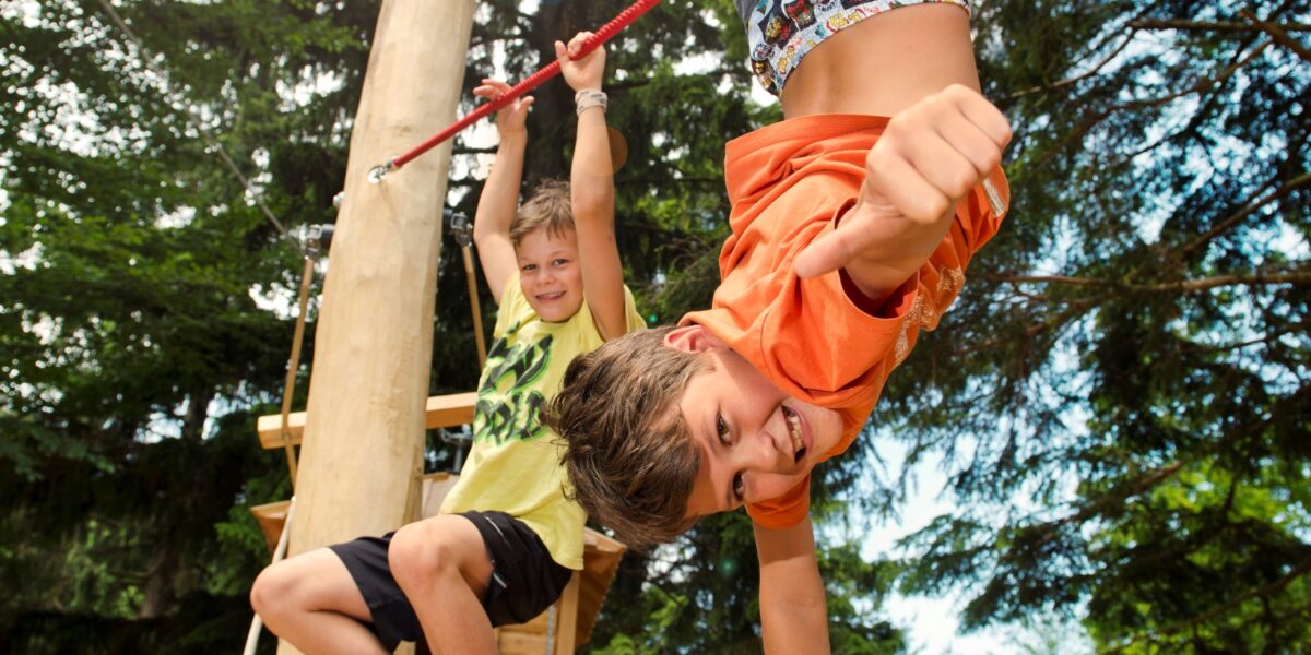 Zwei Kinder spielen auf einem roten Hochseilgarten im Freien. Das eine hängt lächelnd kopfüber, das andere klettert dahinter, umgeben von Bäumen.