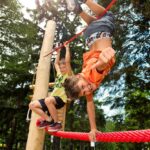 Zwei Kinder spielen auf einem roten Hochseilgarten im Freien. Das eine hängt lächelnd kopfüber, das andere klettert dahinter, umgeben von Bäumen.