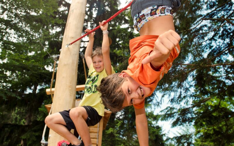 Zwei Kinder spielen auf einem roten Hochseilgarten im Freien. Das eine hängt lächelnd kopfüber, das andere klettert dahinter, umgeben von Bäumen.