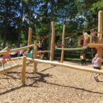 An einem sonnigen Tag spielen Kinder auf einem hölzernen Spielplatzgerüst mit Seilen und Kletternetzen, umgeben von Bäumen.