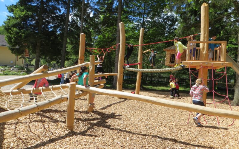An einem sonnigen Tag spielen Kinder auf einem hölzernen Spielplatzgerüst mit Seilen und Kletternetzen, umgeben von Bäumen.