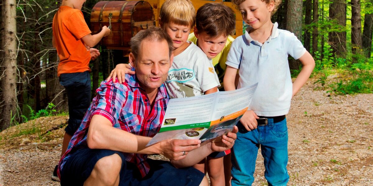 Ein Erwachsener kniet auf einem Waldweg, hält eine Karte in der Hand und spricht mit drei Jungen, während ein anderes Kind in einem orangefarbenen Hemd neben einem Schild und einer Truhe im Hintergrund steht.