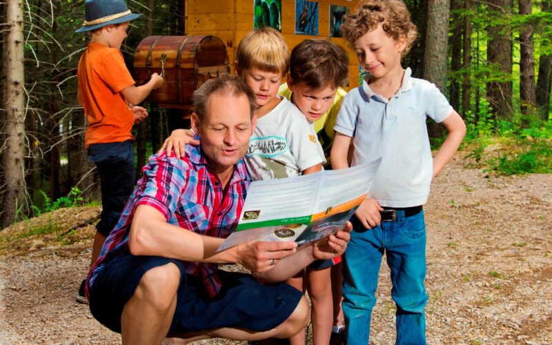 Ein Erwachsener kniet auf einem Waldweg, hält eine Karte in der Hand und spricht mit drei Jungen, während ein anderes Kind in einem orangefarbenen Hemd neben einem Schild und einer Truhe im Hintergrund steht.
