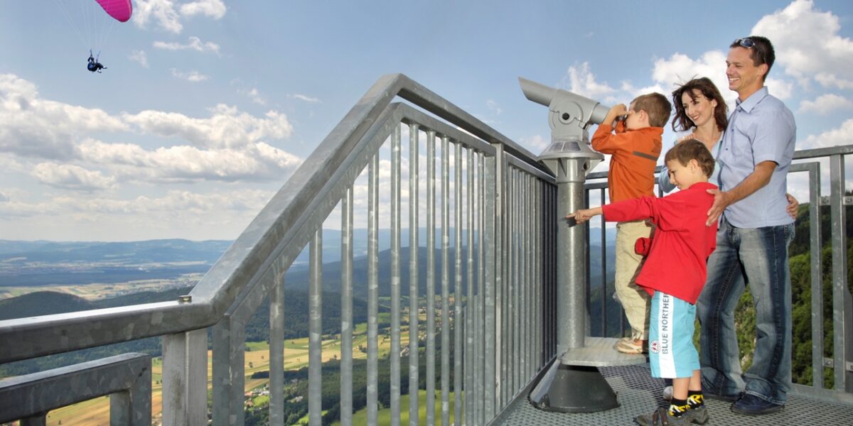 Eine Familie steht auf einer Aussichtsplattform mit Blick auf ein Tal; ein Kind benutzt ein Teleskop, während im Hintergrund ein Gleitschirm am Himmel fliegt.