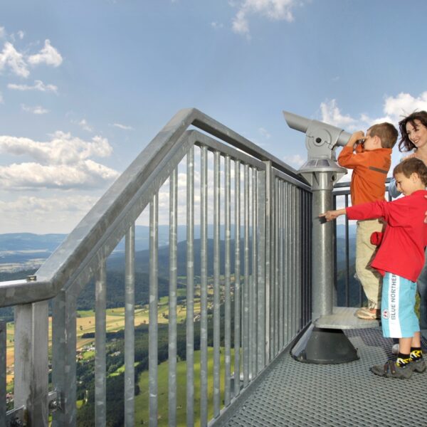 Eine Familie steht auf einer Aussichtsplattform mit Blick auf ein Tal; ein Kind benutzt ein Teleskop, während im Hintergrund ein Gleitschirm am Himmel fliegt.