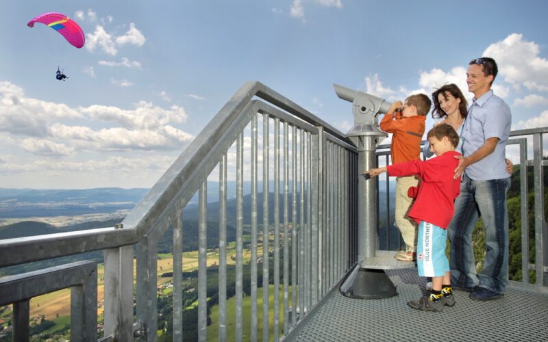 Eine Familie steht auf einer Aussichtsplattform mit Blick auf ein Tal; ein Kind benutzt ein Teleskop, während im Hintergrund ein Gleitschirm am Himmel fliegt.