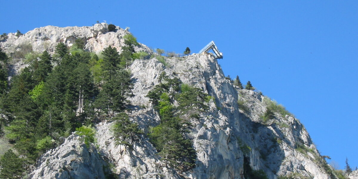 Eine Skisprungschanze befindet sich am Rande eines felsigen, baumbestandenen Berges unter strahlend blauem Himmel.
