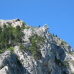 Eine Skisprungschanze befindet sich am Rande eines felsigen, baumbestandenen Berges unter strahlend blauem Himmel.