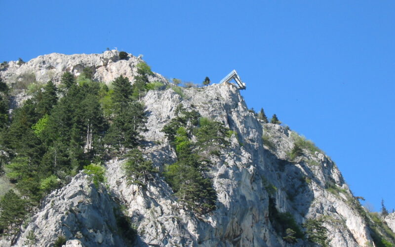 Eine Skisprungschanze befindet sich am Rande eines felsigen, baumbestandenen Berges unter strahlend blauem Himmel.