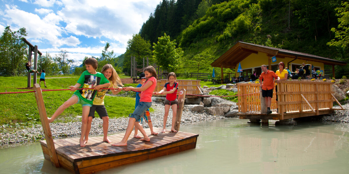 Kinder ziehen sich an einem Seil über einen flachen Teich auf einer hölzernen Plattform, während Erwachsene von einer nahe gelegenen Brücke in einem grünen Park zusehen.