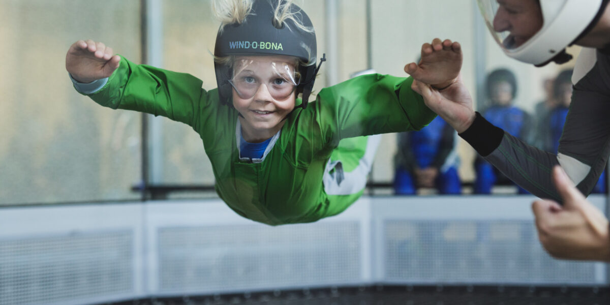 Ein Kind in einem grünen Anzug und Helm schwebt in einem vertikalen Windkanal, während ein Lehrer seinen Arm zur Anleitung hält.