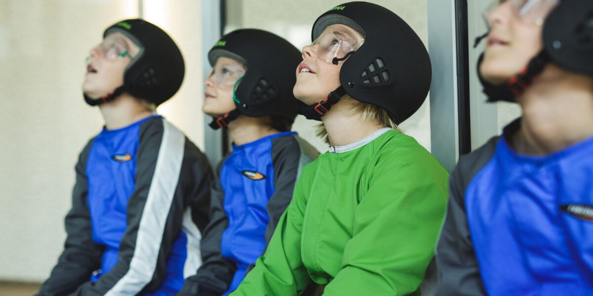 Vier Kinder mit Helmen, Overalls und Schutzbrillen sitzen drinnen und schauen mit aufmerksamen Blicken nach oben.