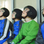 Vier Kinder mit Helmen, Overalls und Schutzbrillen sitzen drinnen und schauen mit aufmerksamen Blicken nach oben.