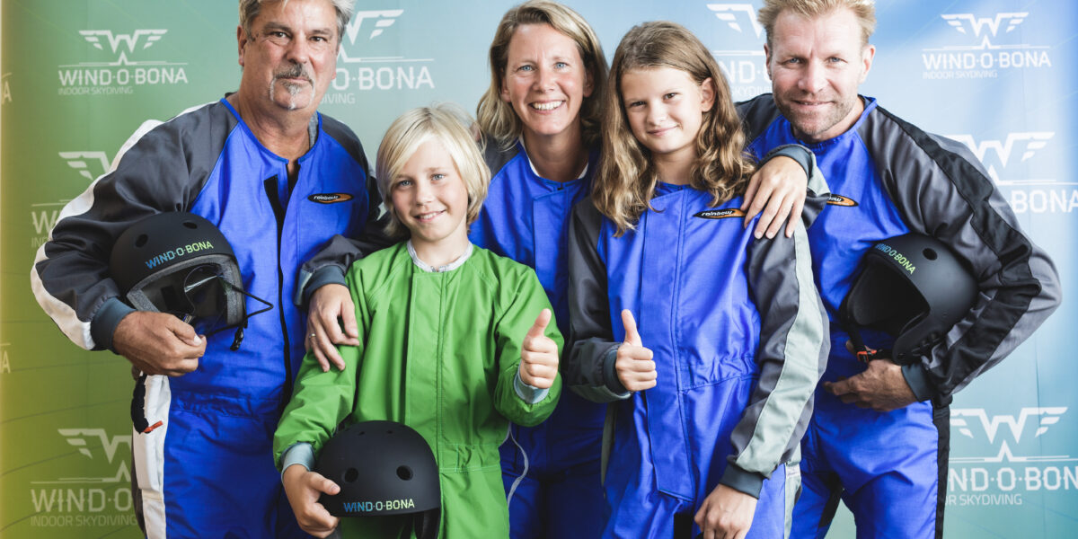Fünf Personen in Fluganzügen posieren lächelnd und mit erhobenem Daumen für ein Gruppenfoto vor einer Indoor-Skydiving-Kulisse von Windobona.