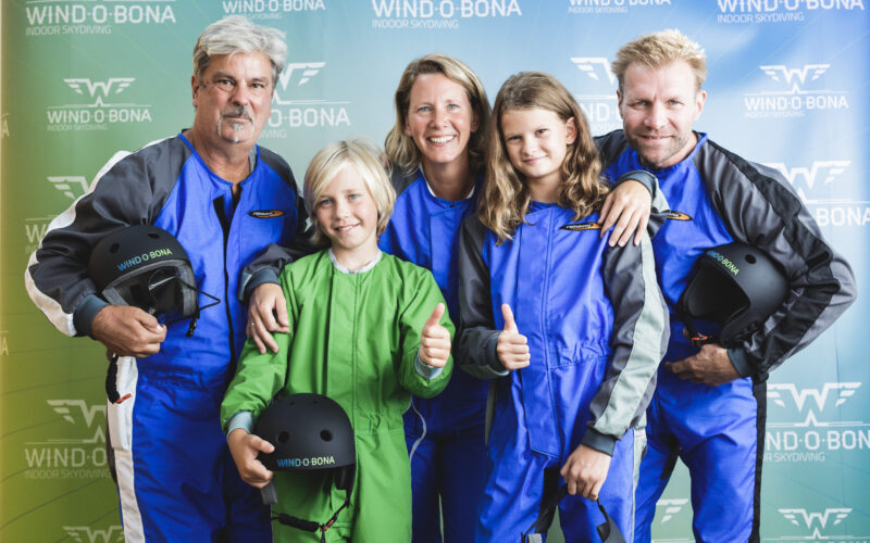 Fünf Personen in Fluganzügen posieren lächelnd und mit erhobenem Daumen für ein Gruppenfoto vor einer Indoor-Skydiving-Kulisse von Windobona.