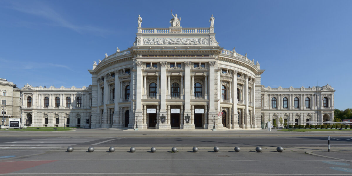 Frontansicht eines großen neoklassizistischen Gebäudes mit Säulen, Statuen auf dem Dach und kunstvollen architektonischen Details vor einem klaren blauen Himmel.