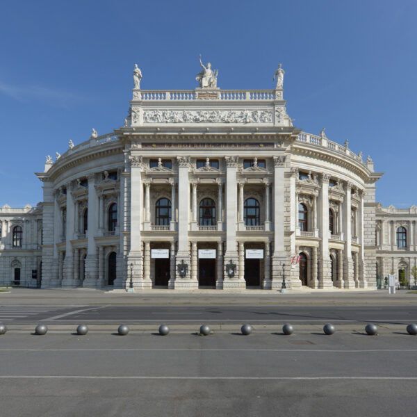 Frontansicht eines großen neoklassizistischen Gebäudes mit Säulen, Statuen auf dem Dach und kunstvollen architektonischen Details vor einem klaren blauen Himmel.