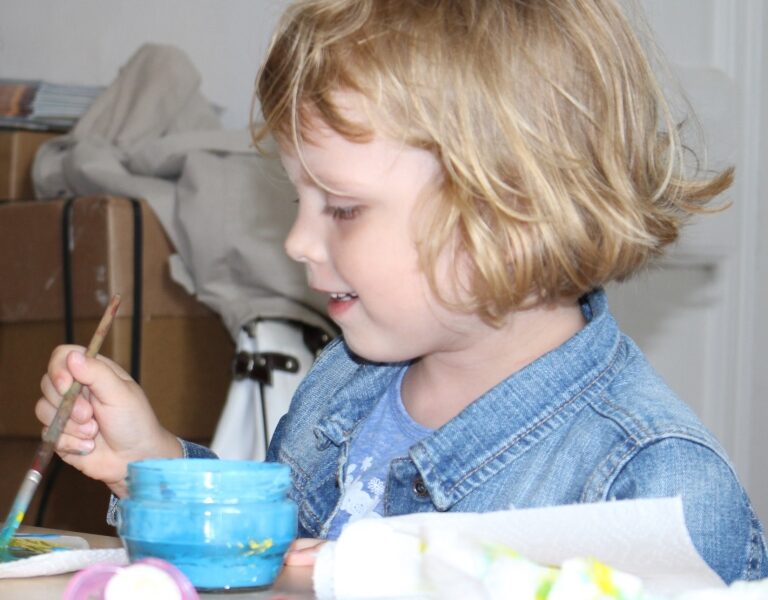 Ein Kind mit blondem Haar und Jeansjacke malt mit einem Pinsel und blauer Farbe aus einem Glas, während es an einem mit Papiertüchern und Malutensilien bedeckten Tisch sitzt.