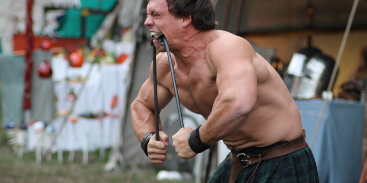 Ein hemdsärmeliger Mann im Kilt verbiegt bei einer Veranstaltung oder Performance im Freien eine Metallstange mit Mund und Händen.