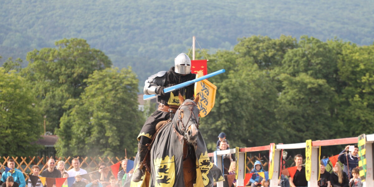 Ein Ritter in Rüstung reitet auf einem Pferd und hält eine blaue Lanze während eines Ritterturniers, mit Zuschauern und einem baumbestandenen Hügel im Hintergrund.