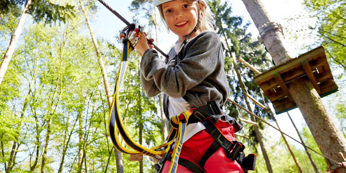 Ein Kind mit Helm und Klettergurt hält sich in einem Hochseilgarten im Wald an Seilen fest.
