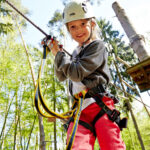 Ein Kind mit Helm und Klettergurt hält sich in einem Hochseilgarten im Wald an Seilen fest.