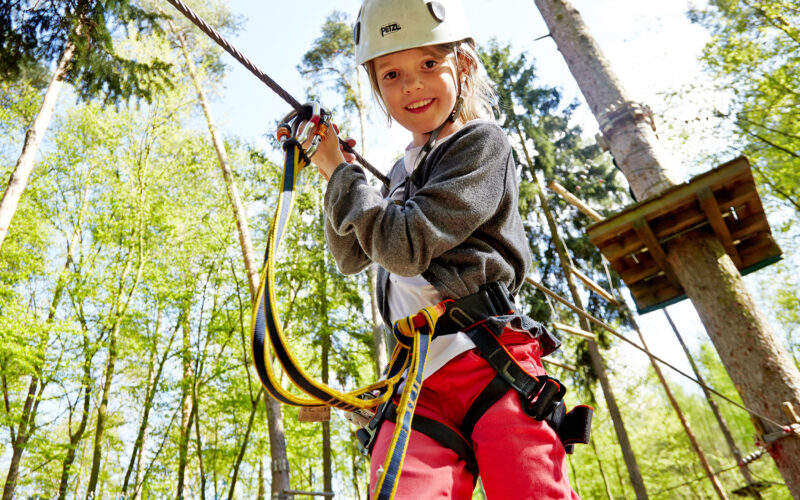 Ein Kind mit Helm und Klettergurt hält sich in einem Hochseilgarten im Wald an Seilen fest.