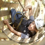 Ein Kind, das einen Helm und einen Sicherheitsgurt trägt, lächelt beim Klettern durch einen Holztunnel in einem Hochseilgarten.